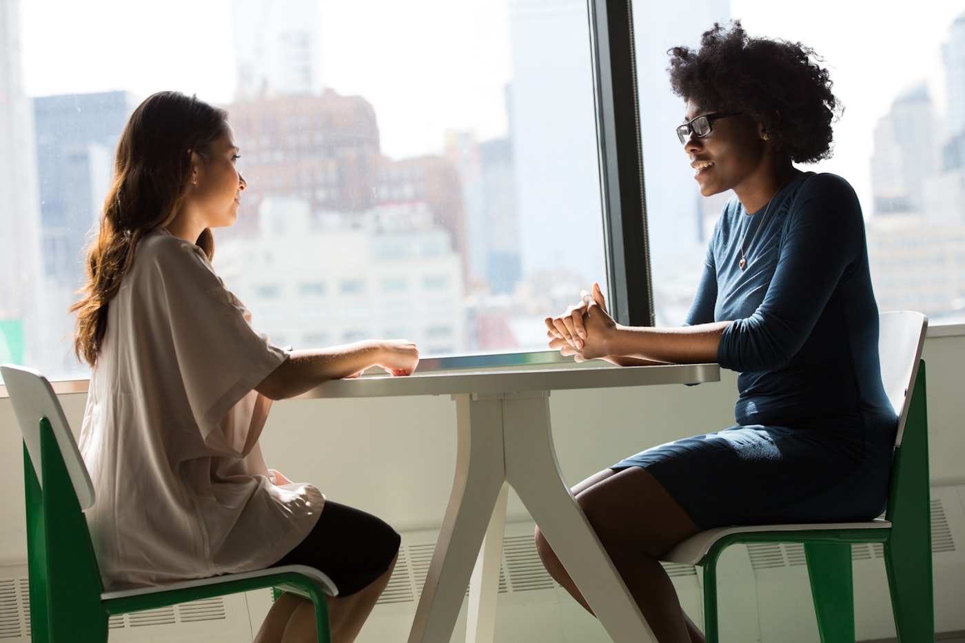 Two women sitting across a table by a window, talking with each other.