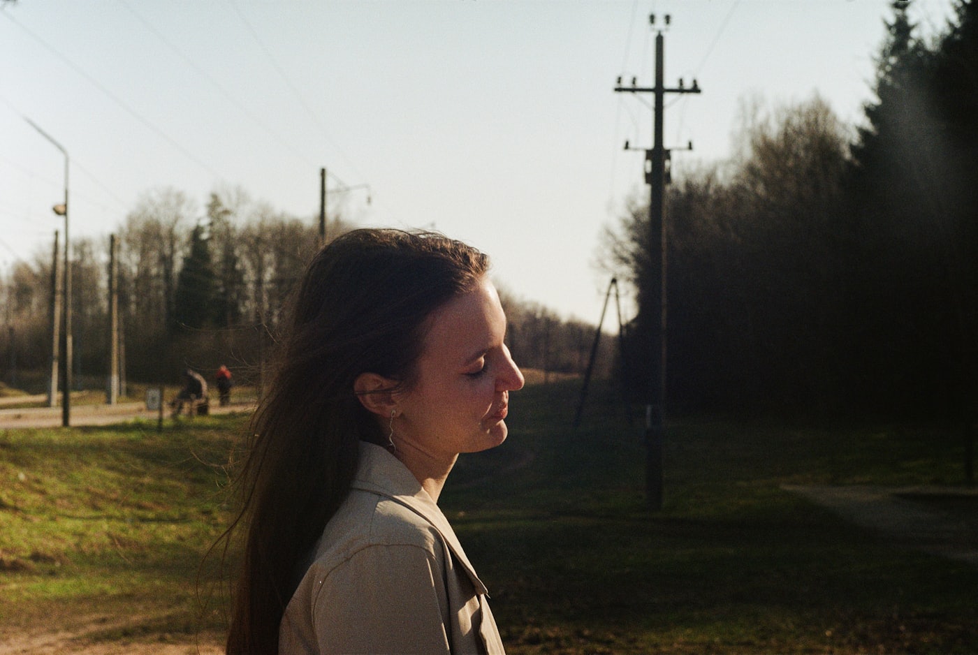 A woman standing outdoors in evening light, looking away in thought.